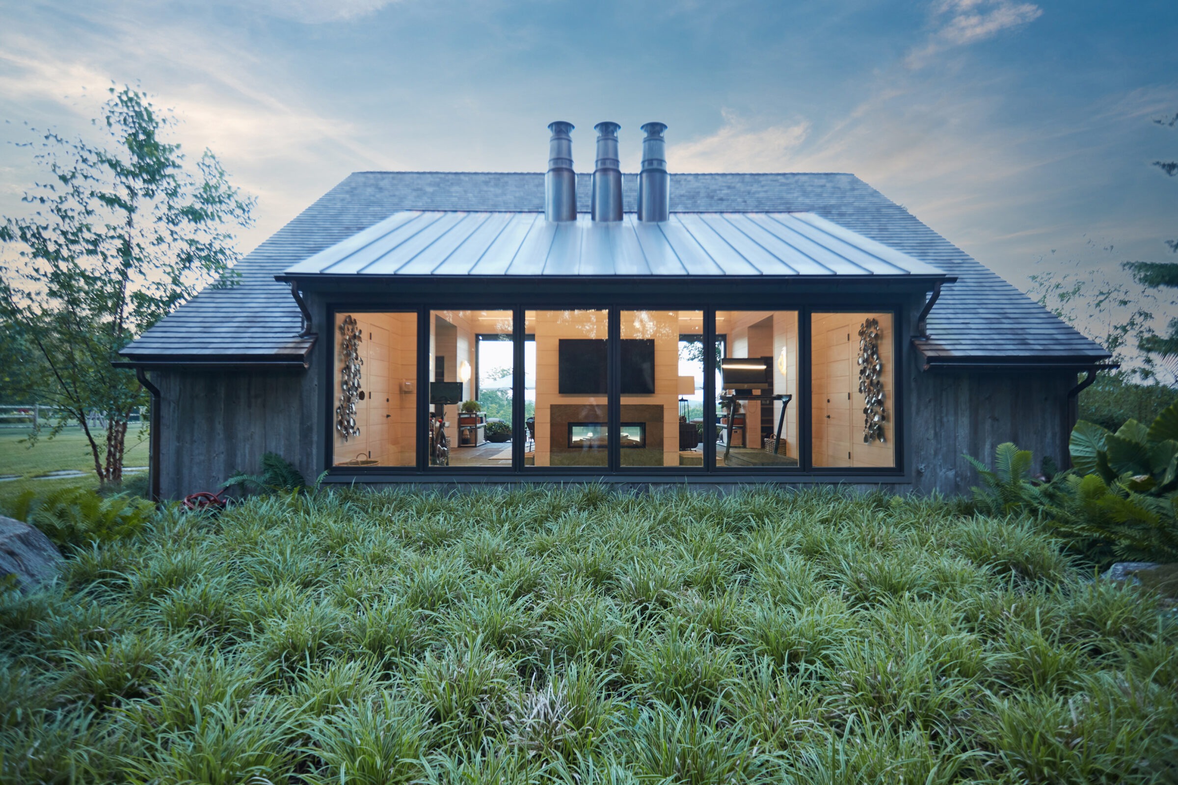 Modern cabin with large windows and metal roof surrounded by lush grass. The exterior is dark wood, with a clear view inside. Three chimneys visible.
