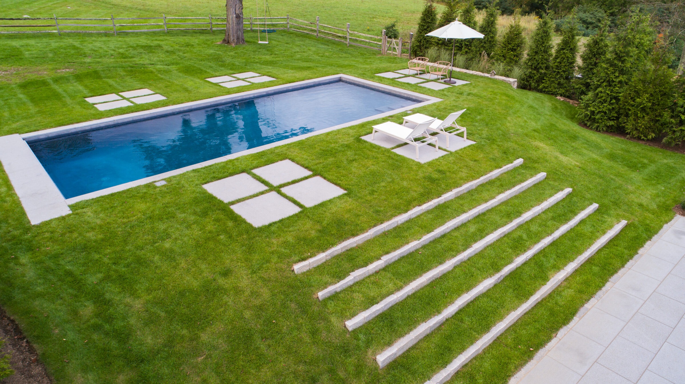 An aerial view of a backyard featuring a rectangular pool, loungers, green lawn, stepping stones, wood rail fence, and a parasol.