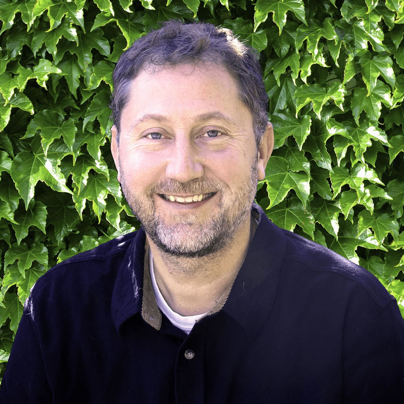A smiling person with a beard poses in front of a lush ivy background. They are wearing a dark collar shirt and seem content.