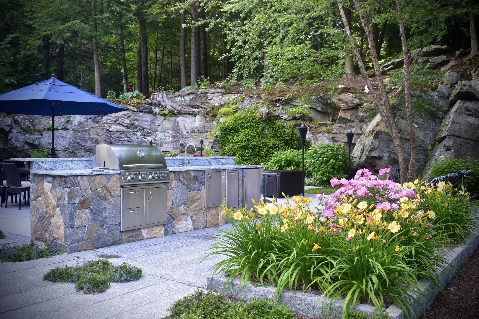 An outdoor kitchen with a built-in grill and countertop set against a backdrop of a natural rock formation, surrounded by a garden with colorful flowers.