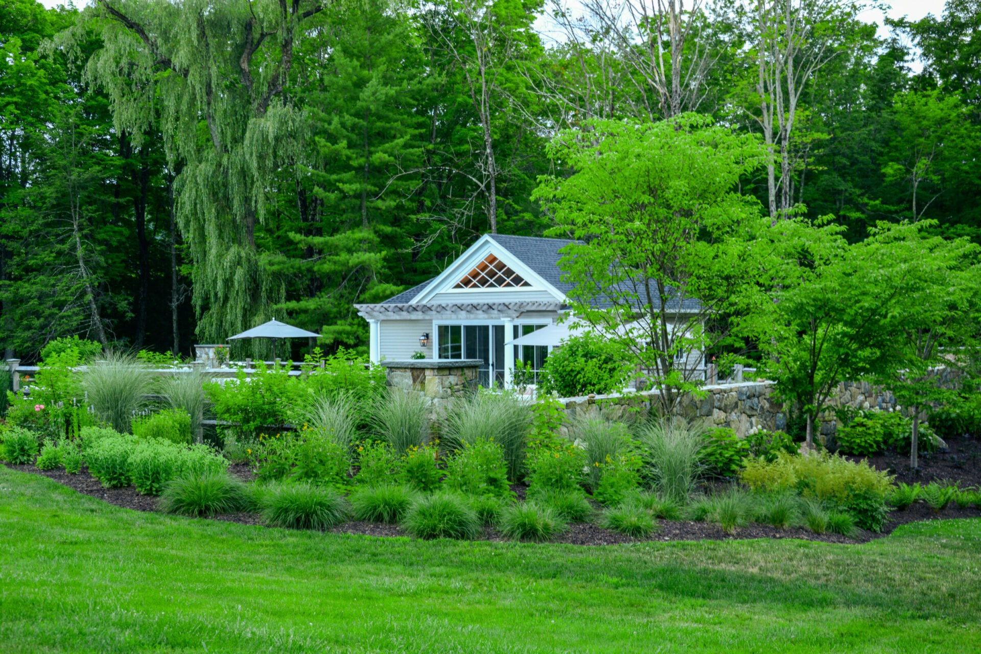 A quaint white building surrounded by lush greenery, trees, and neatly landscaped gardens, with an outdoor umbrella visible in the background.