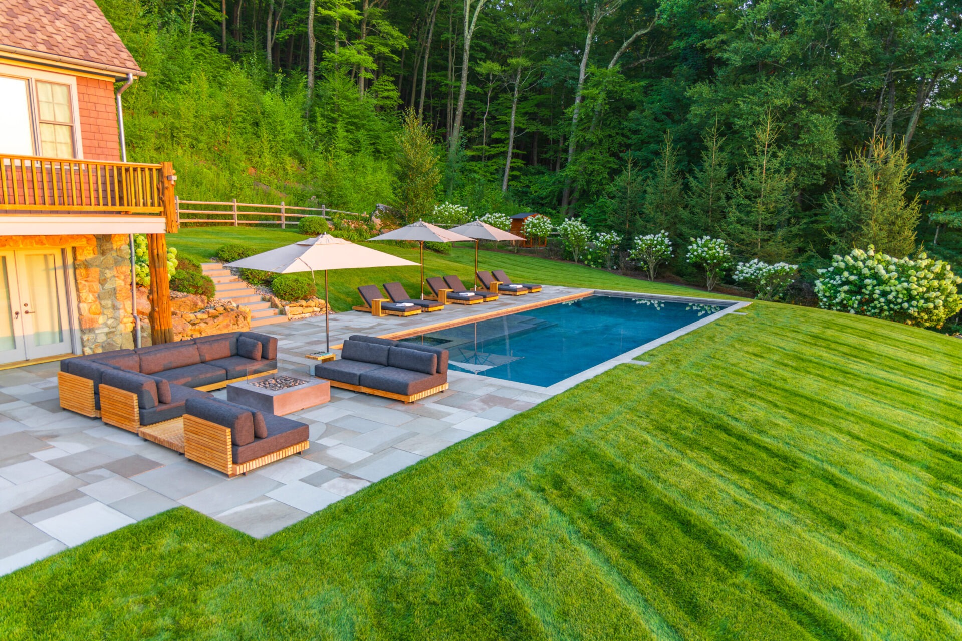 A luxurious backyard with an in-ground pool, trimmed lawn, patio furniture, and large umbrellas adjacent to a house with a wooded backdrop.