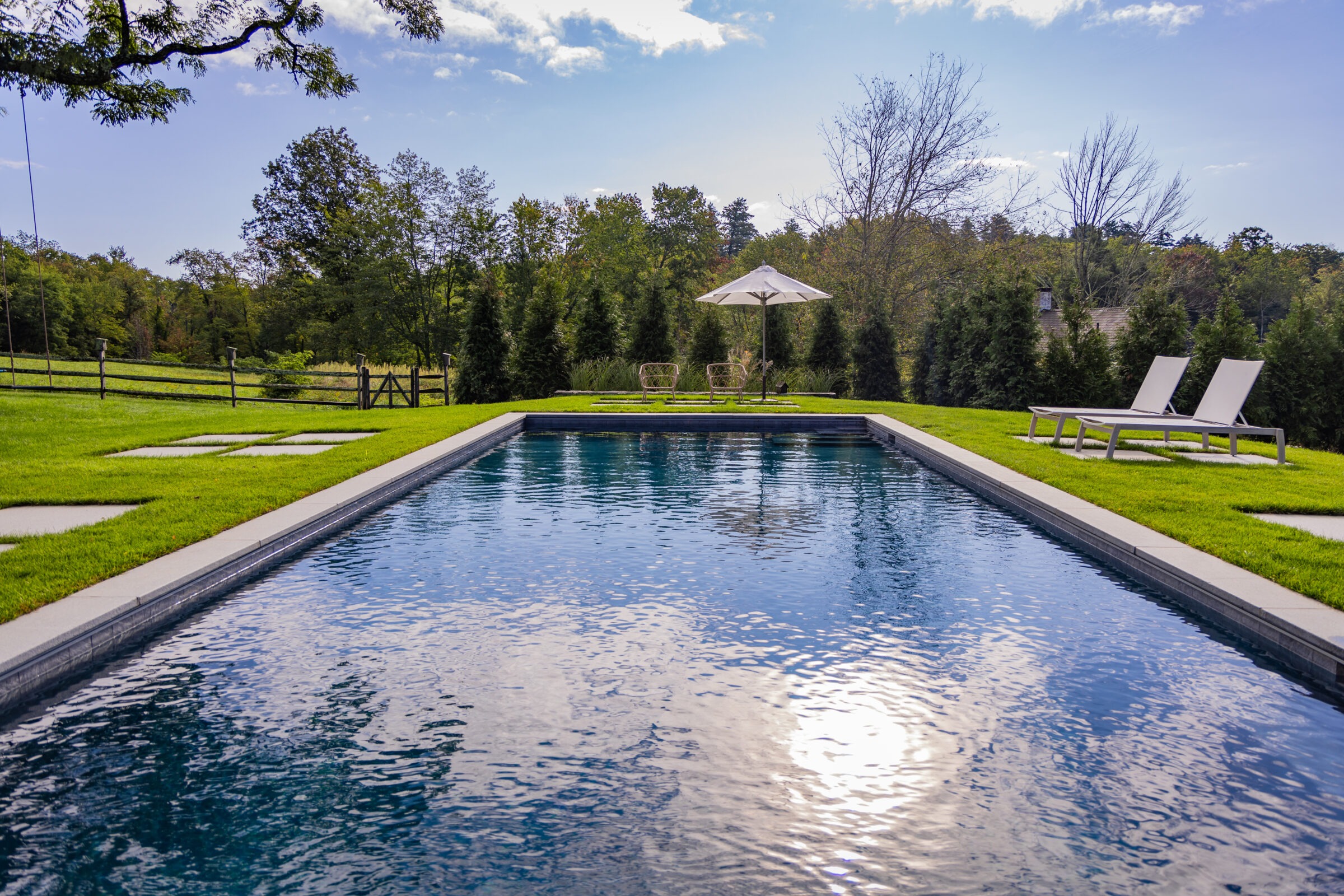 A serene outdoor swimming pool with loungers and an umbrella, surrounded by a well-manicured lawn, trees, and a clear blue sky.