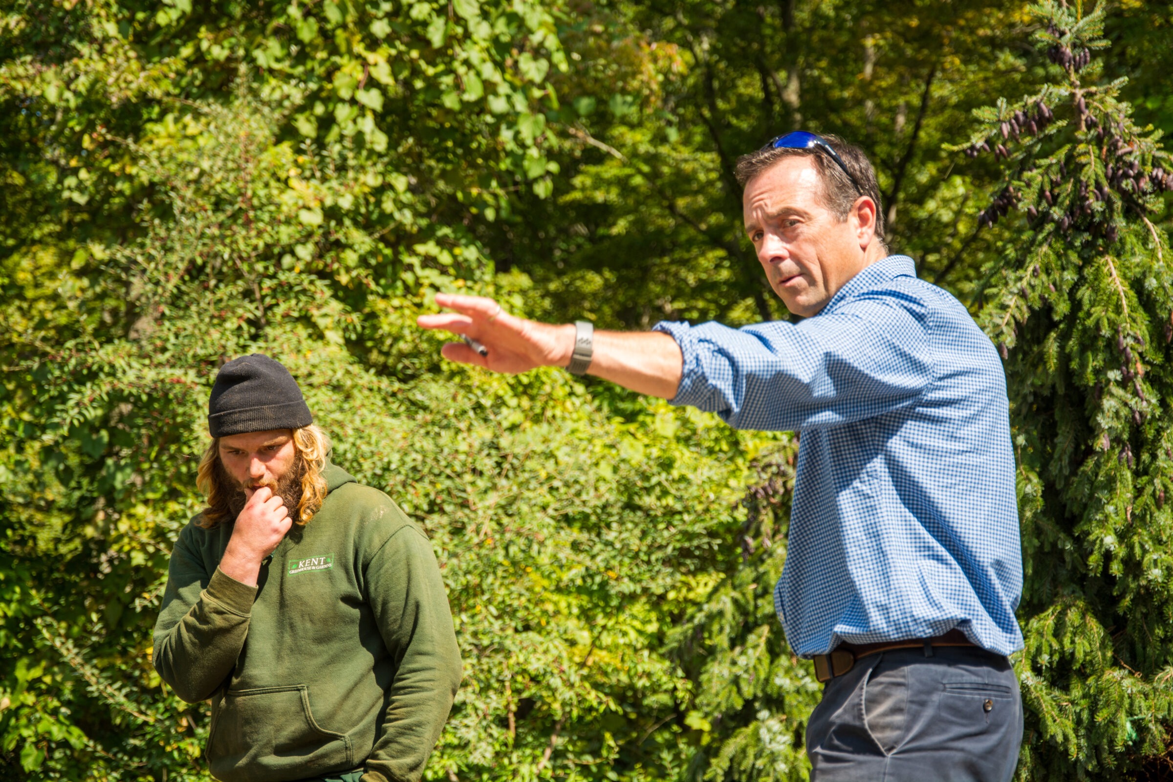 One person is gesturing and speaking while another listens intently outdoors, surrounded by lush green foliage on a sunny day.