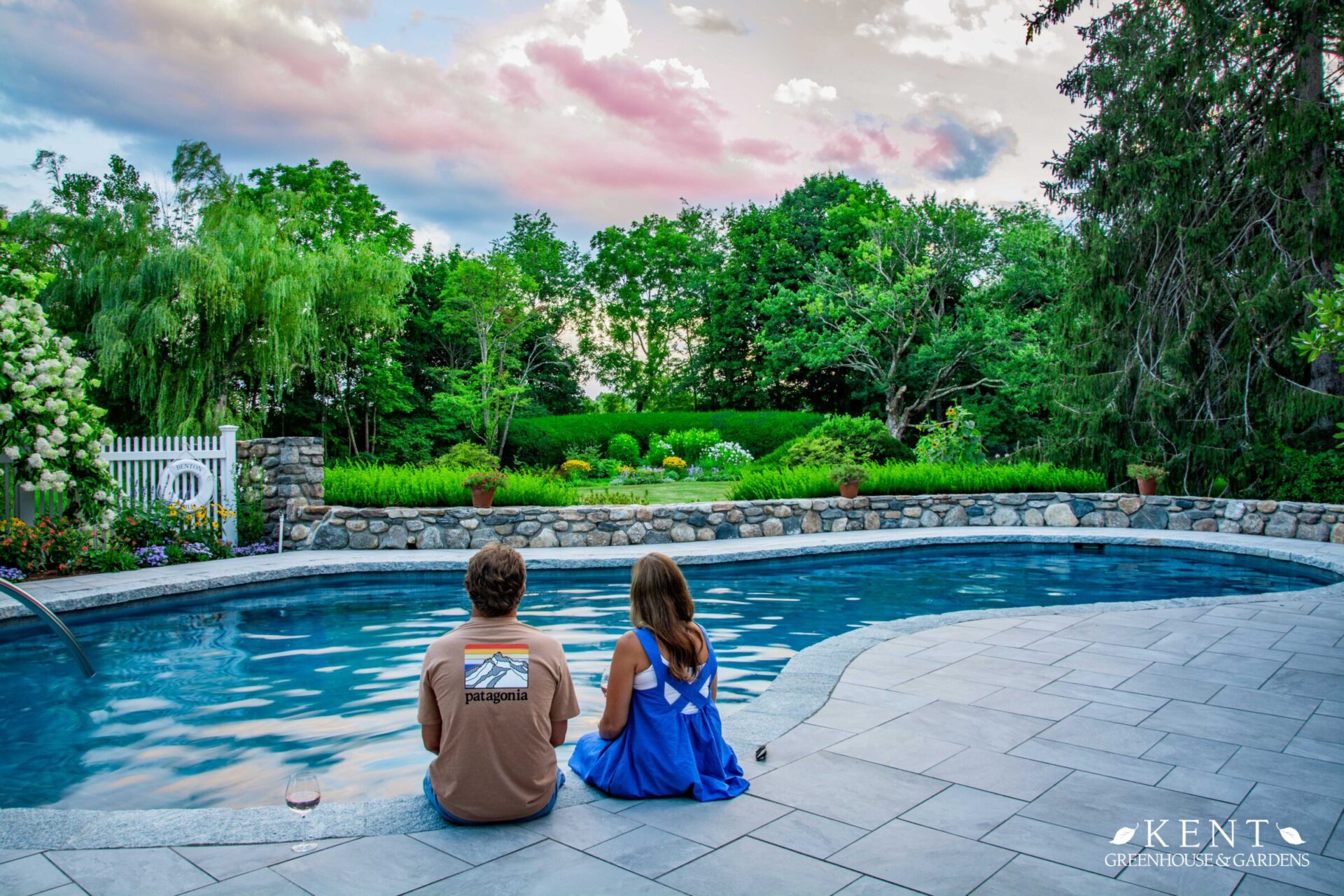 Two people sit by a serene pool, gazing at a lush garden, under a pastel sky at dusk, exuding a tranquil, picturesque summer evening ambiance.