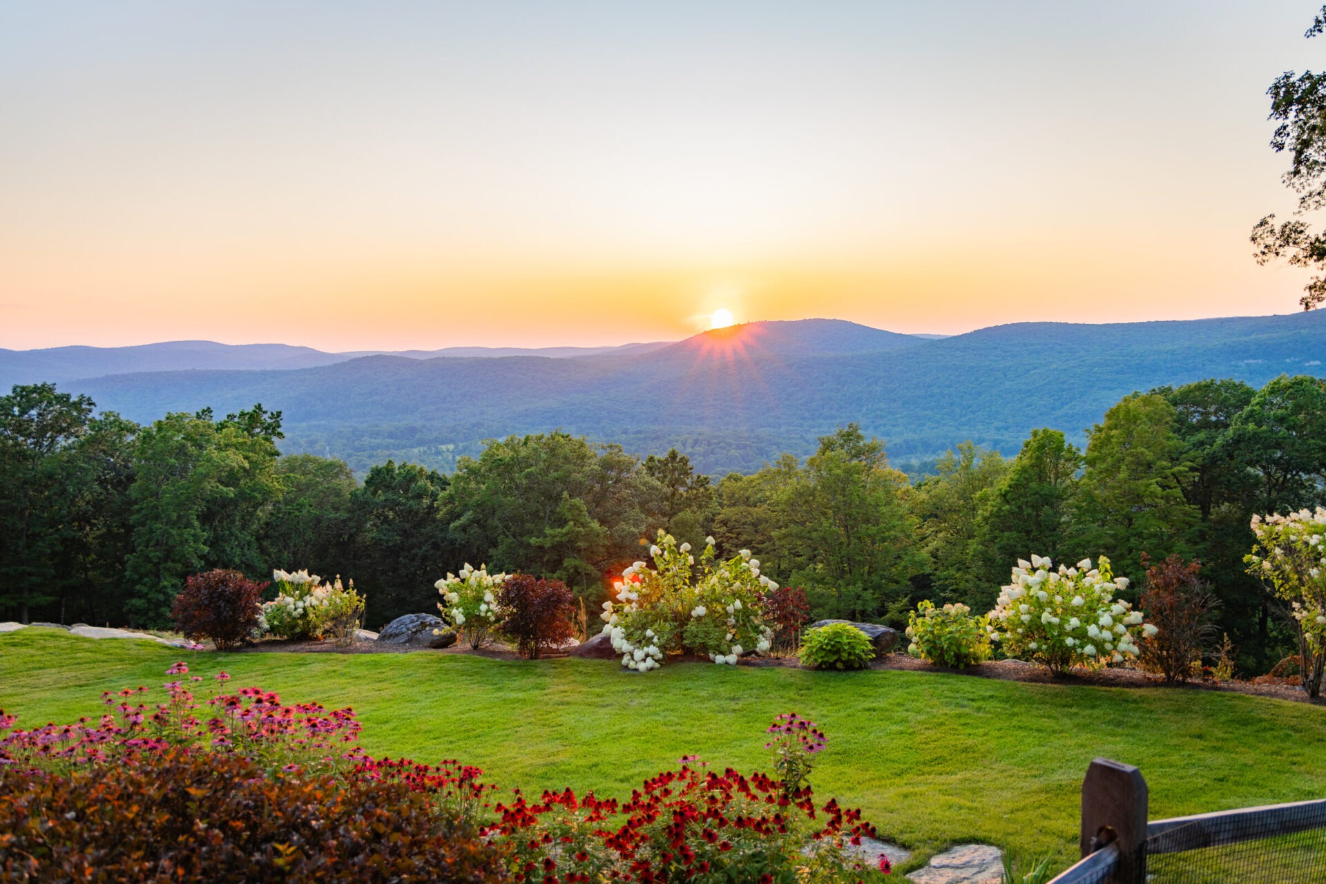A tranquil sunset over rolling hills, viewed from a lush garden adorned with colorful flowers and shrubs, under a softly gradient sky.