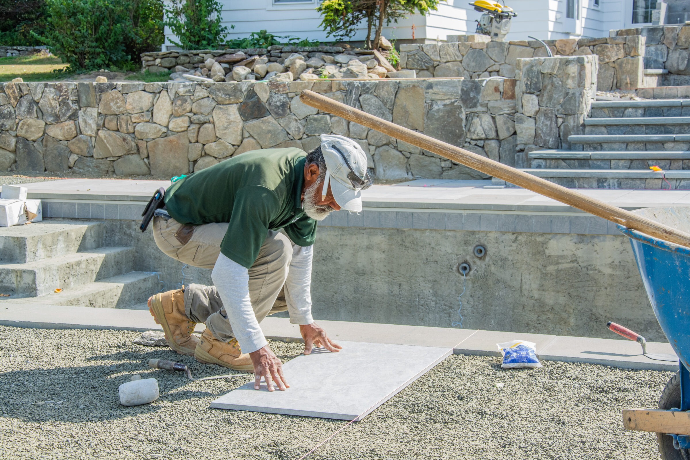 A person is installing a large tile outdoors, kneeling on a substrate with tools nearby, near an unfinished pool and stone steps under sunlight.