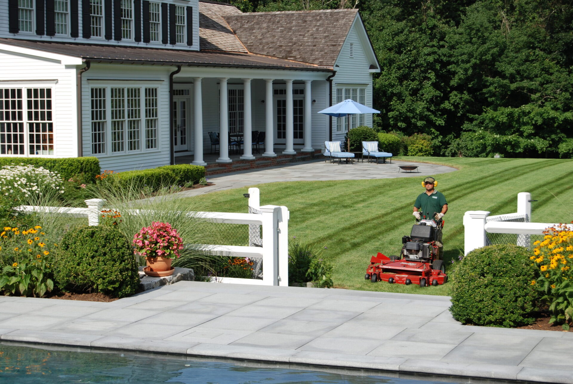 A person is mowing a lawn near a luxurious house with a pool. There are landscaped flowers, white fencing, and lounging chairs in the background.