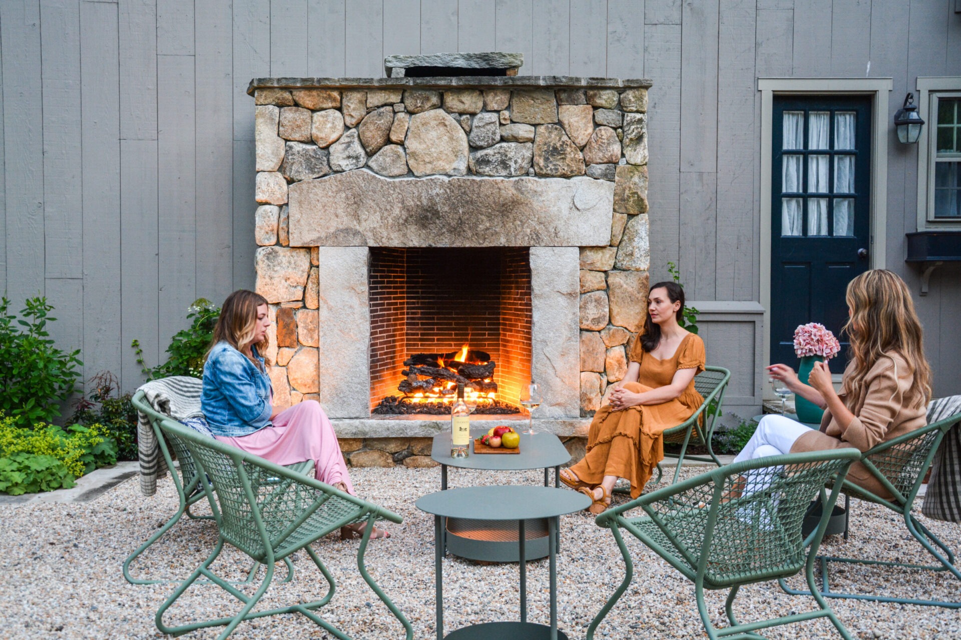 Three people are seated outdoors around a lit fireplace, talking and enjoying each other's company in a cozy, casual backyard setting.