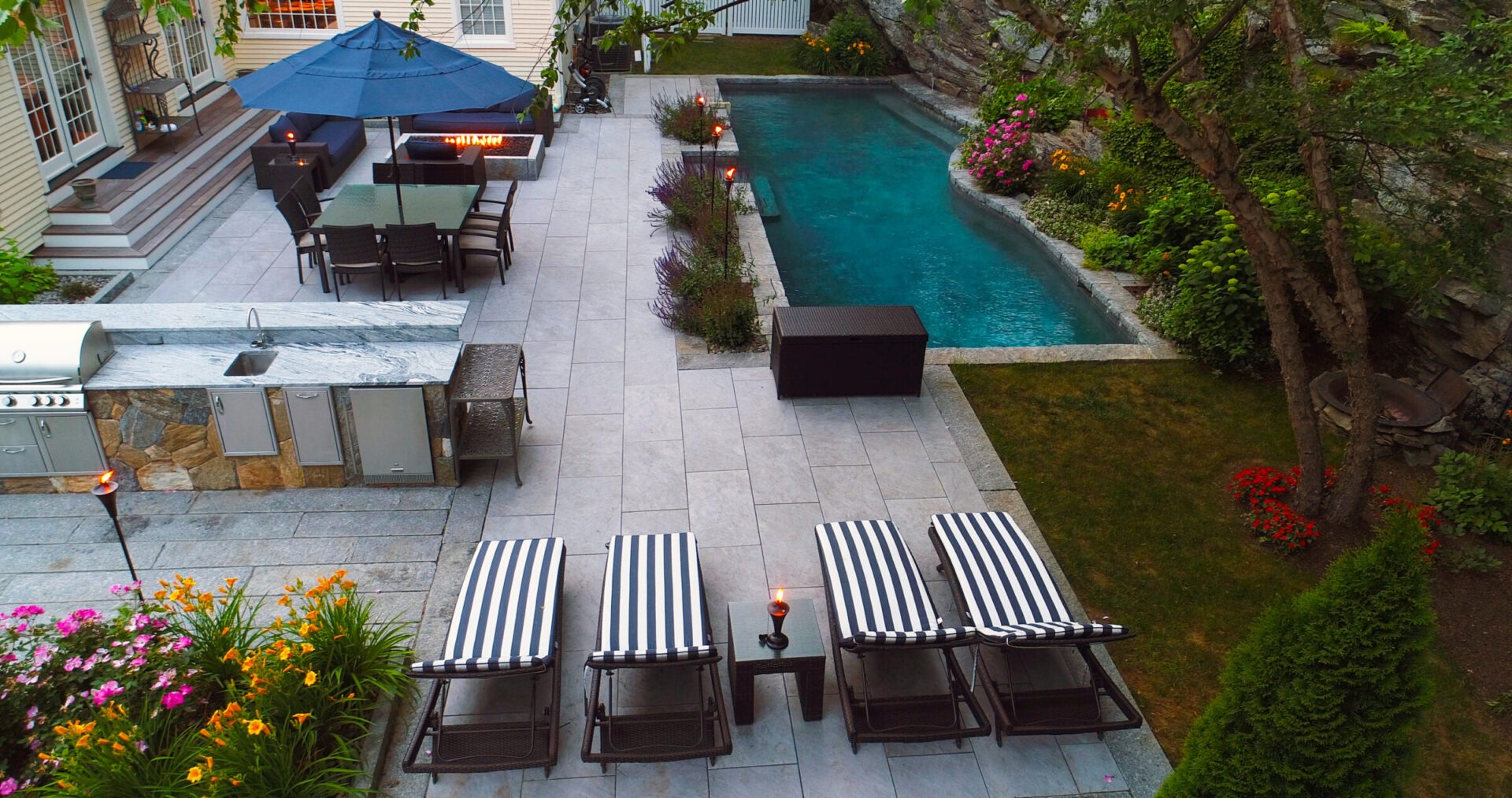 An upscale backyard with a swimming pool, patio, outdoor kitchen, dining area, lounge chairs, vibrant flowers, and lush greenery in a serene setting.
