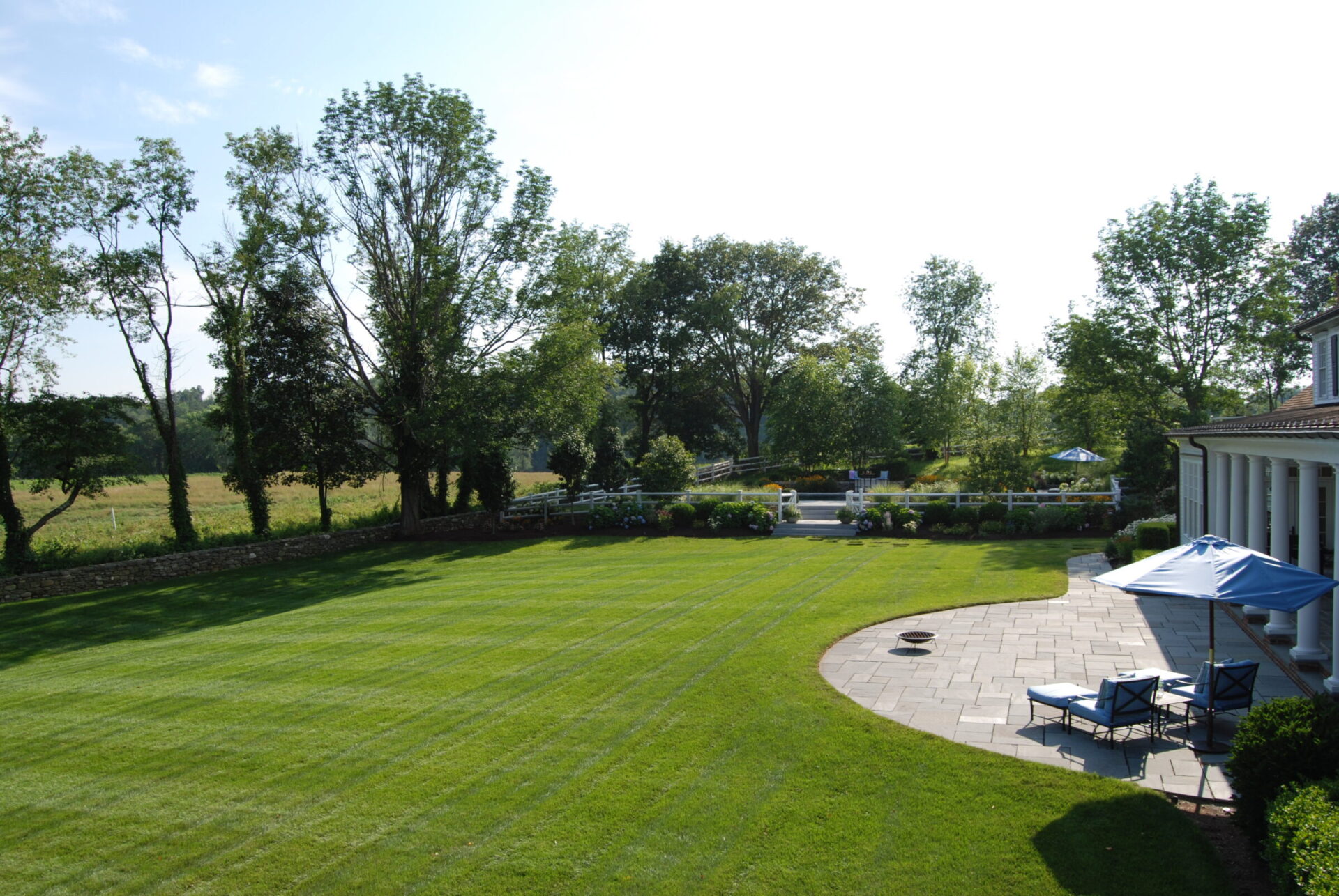The image shows a neatly manicured lawn with striped patterns, a patio area with seating, an umbrella, and a lush landscape with trees under a clear sky.