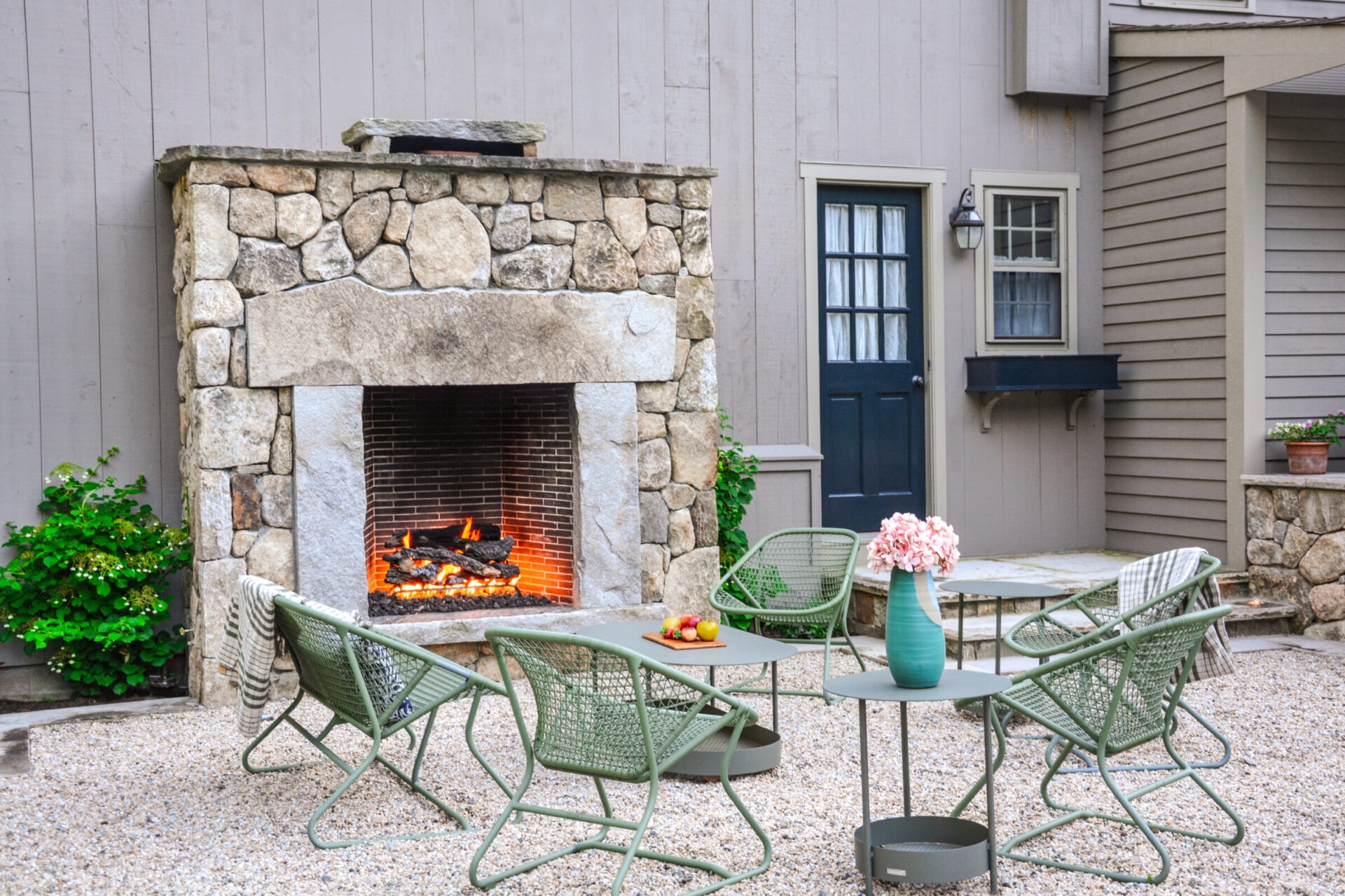 An outdoor patio with an active stone fireplace, green metal chairs, a round table, decorative flowers, and a neutral-colored building with a blue door.