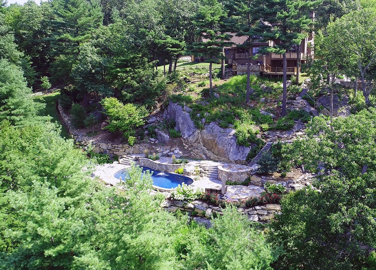 Aerial view of a landscaped property with an irregular-shaped pool amid rocks, trees, and a house with multiple levels, in a serene wooded area.