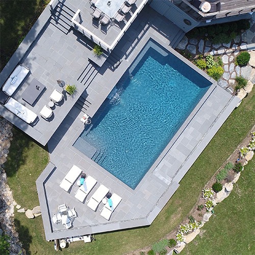 Aerial view of a backyard area with a large, square swimming pool surrounded by loungers, a neat patio, greenery, and part of a house.