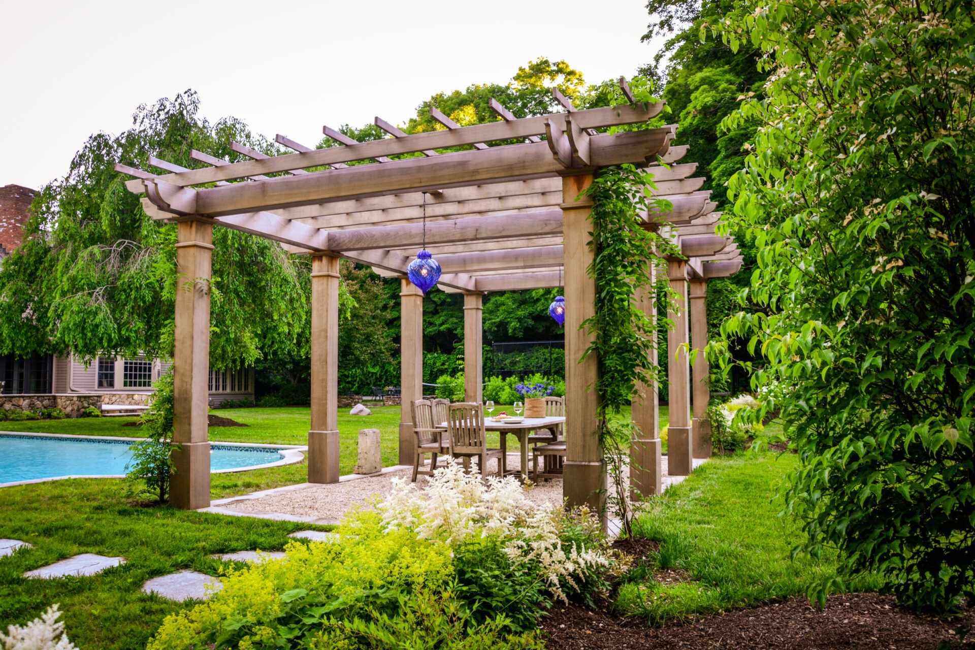 A lush garden setting with a wooden pergola, dining furniture underneath, hanging blue lanterns, a swimming pool in the background, and greenery all around.
