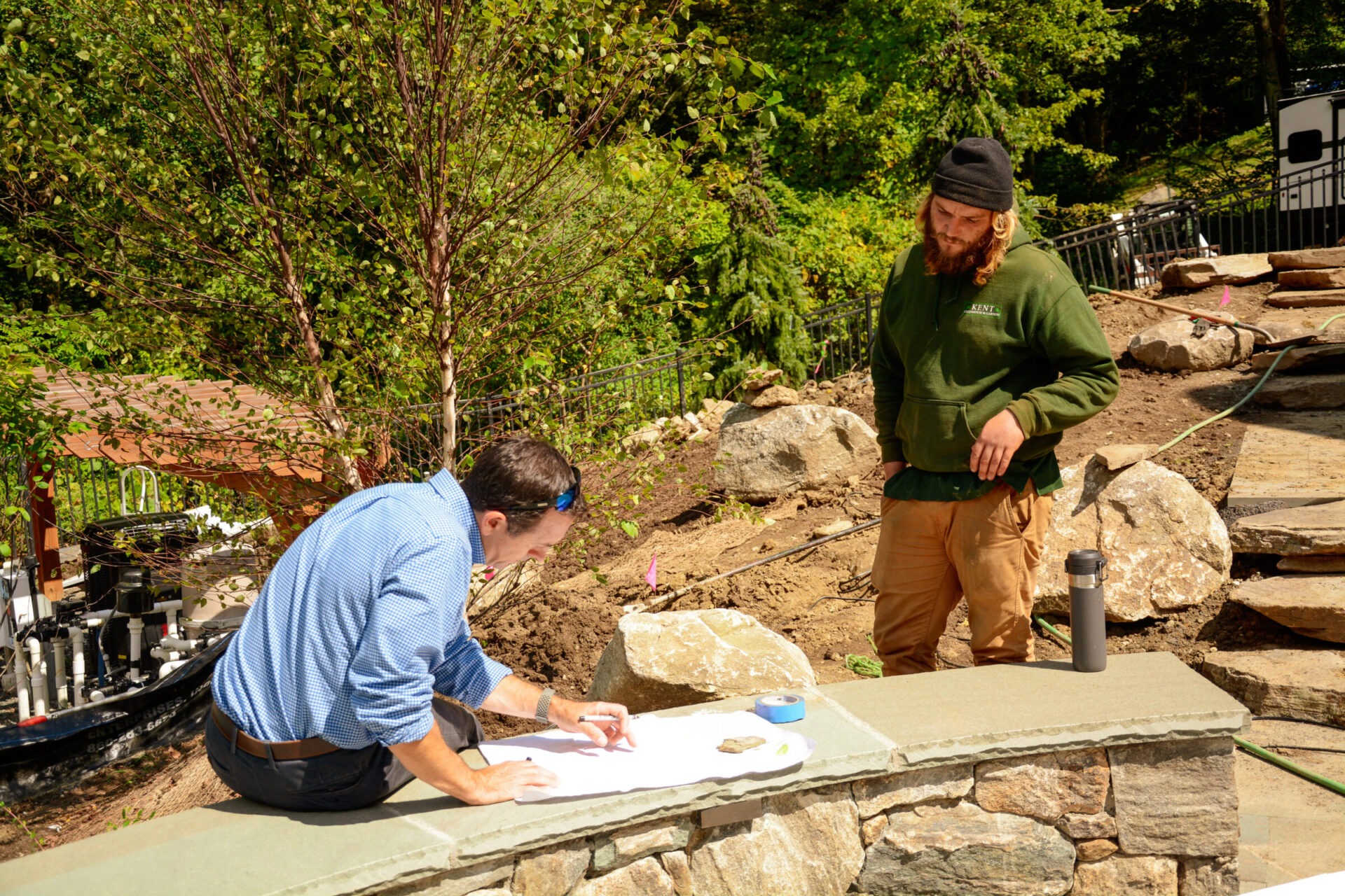 Two people are outdoors; one is reviewing plans on a stone wall while the other observes. They are surrounded by trees, boulders, and garden equipment.