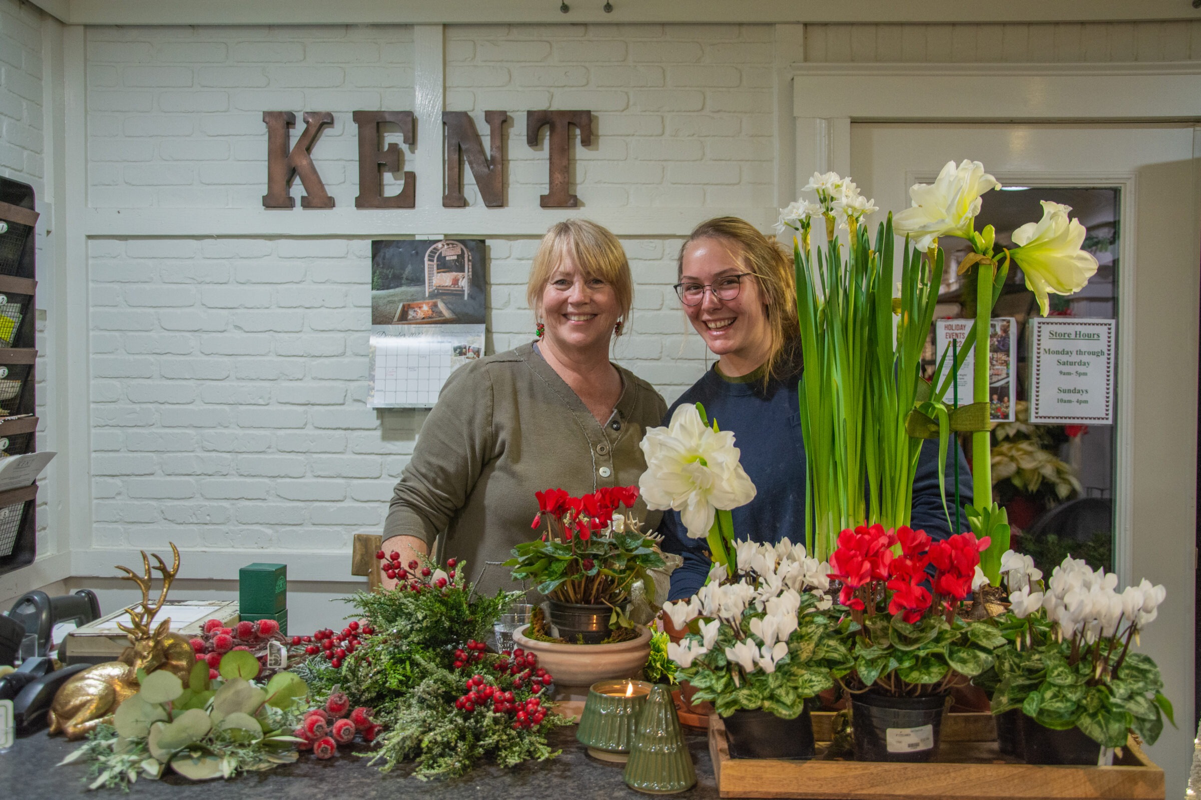 Two people stand smiling behind a counter adorned with vibrant flowers and festive decorations, under a sign that spells "KENT" on a white brick wall.