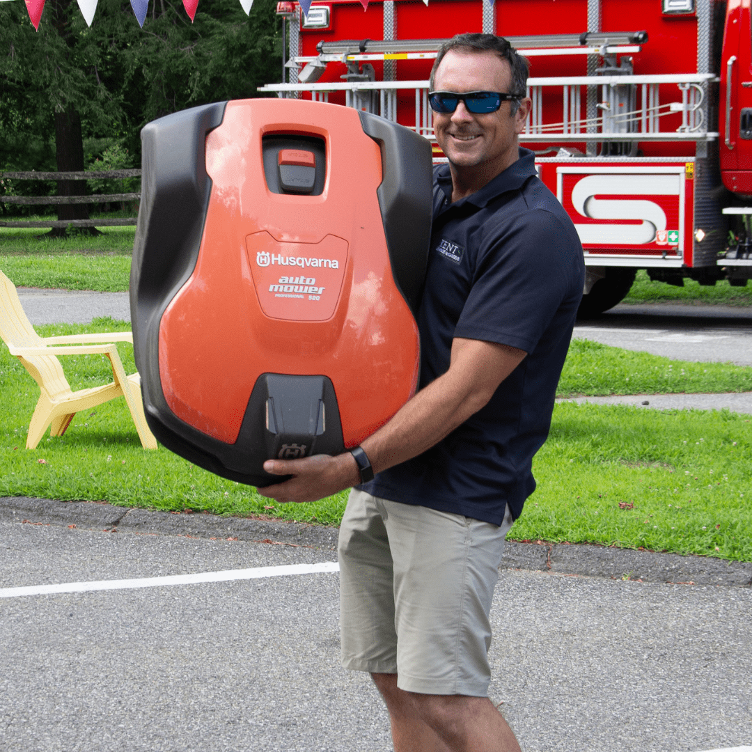 A smiling person is carrying an orange Husqvarna Automower, wearing sunglasses and casual clothing, with a red fire truck and chair in the background.